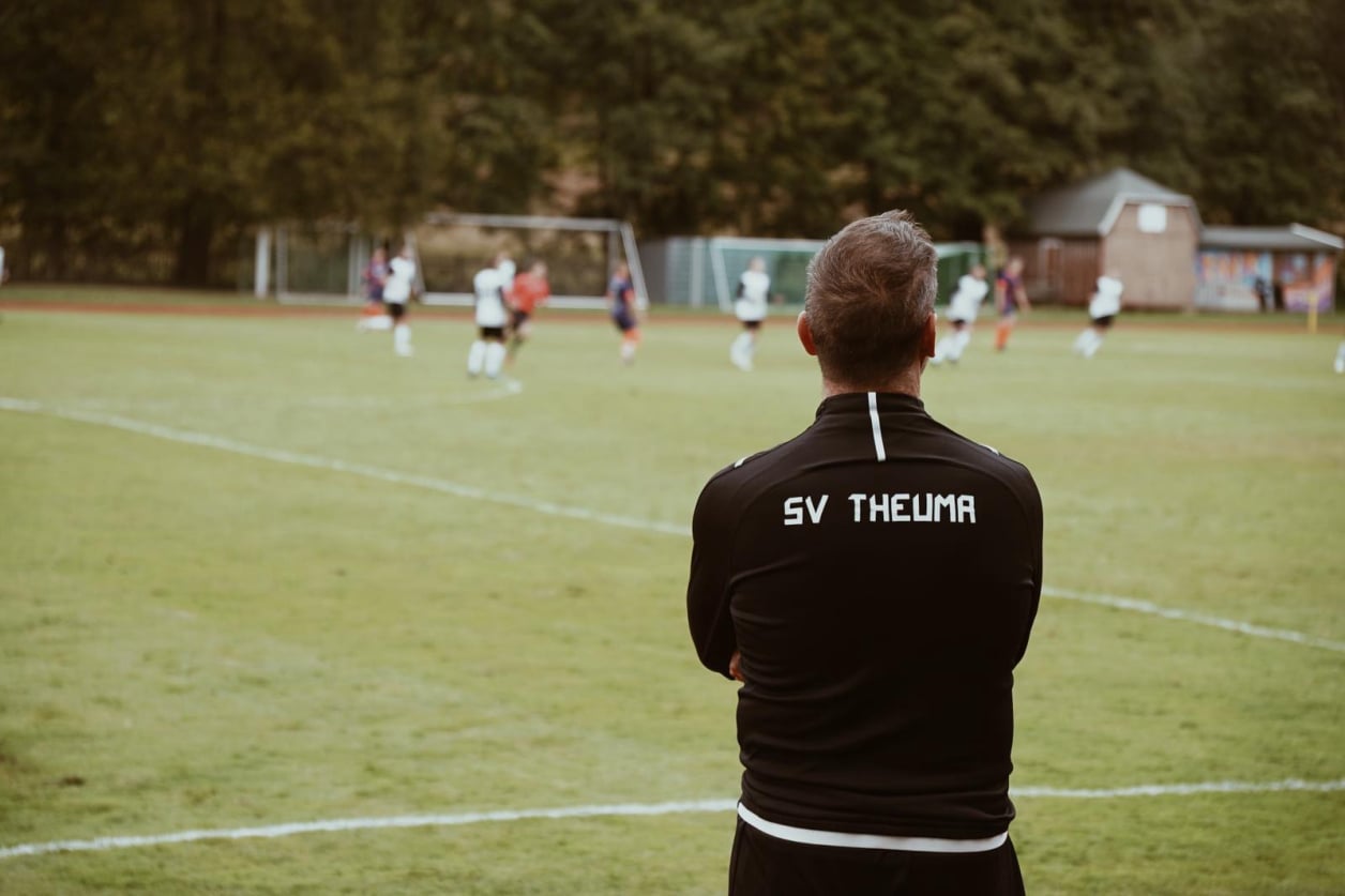 Ein Fuballtrainer von hinten mit Blick auf das Spielfeld, im Hintergrund läuft ein Fußballspiel des SV Theumas