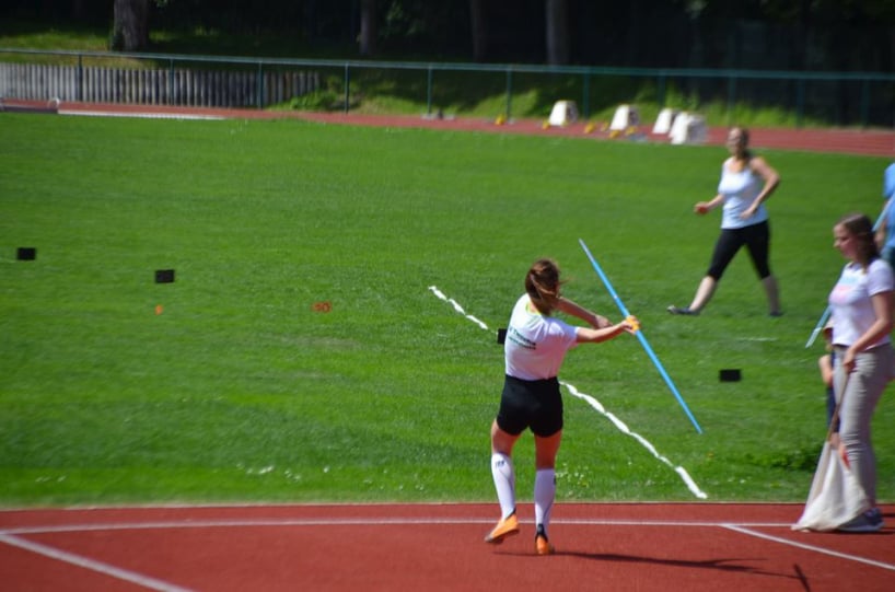 Eine Leichtathletin (von hinten dargestellt) wirft einen Speer - am Rand weitere Personen